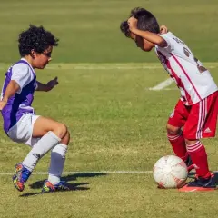 Due ragazzini si sfidano su un campo da calcio in erba, uno in maglia viola, l'altro in maglia bianca e rossa, con un pallone ai loro piedi.