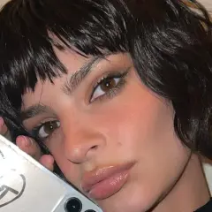 Primo piano di una donna con capelli scuri e umidi che le incorniciano il viso. Tiene in mano un telefono con un disegno a cuore e la scritta "CUNT".
