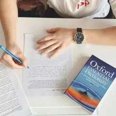 Studente che scrive, con un dizionario Oxford Essential Dictionary aperto. Cosa vuol dire AT? Forse "at" è un'abbreviazione usata in questo contesto di studio.