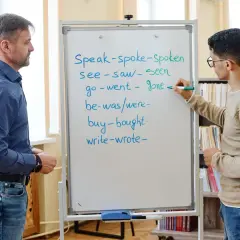 Un insegnante e uno studente studiano il passato in inglese. Lo studente scrive verbi irregolari sulla lavagna.