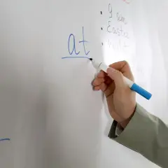 Mano scrive "at" con pennarello blu su lavagna bianca. Lezione di inglese in corso.