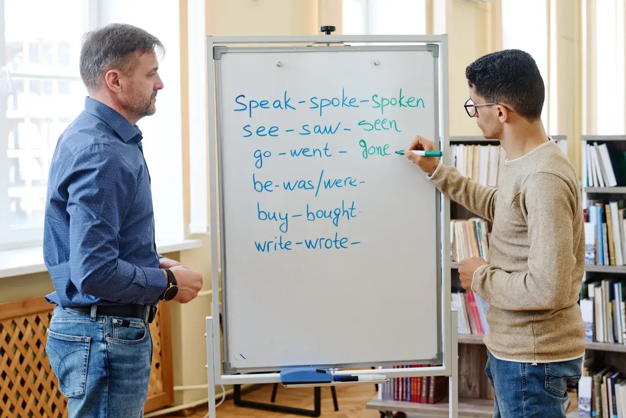 Un insegnante e uno studente studiano il passato in inglese. Lo studente scrive verbi irregolari sulla lavagna.